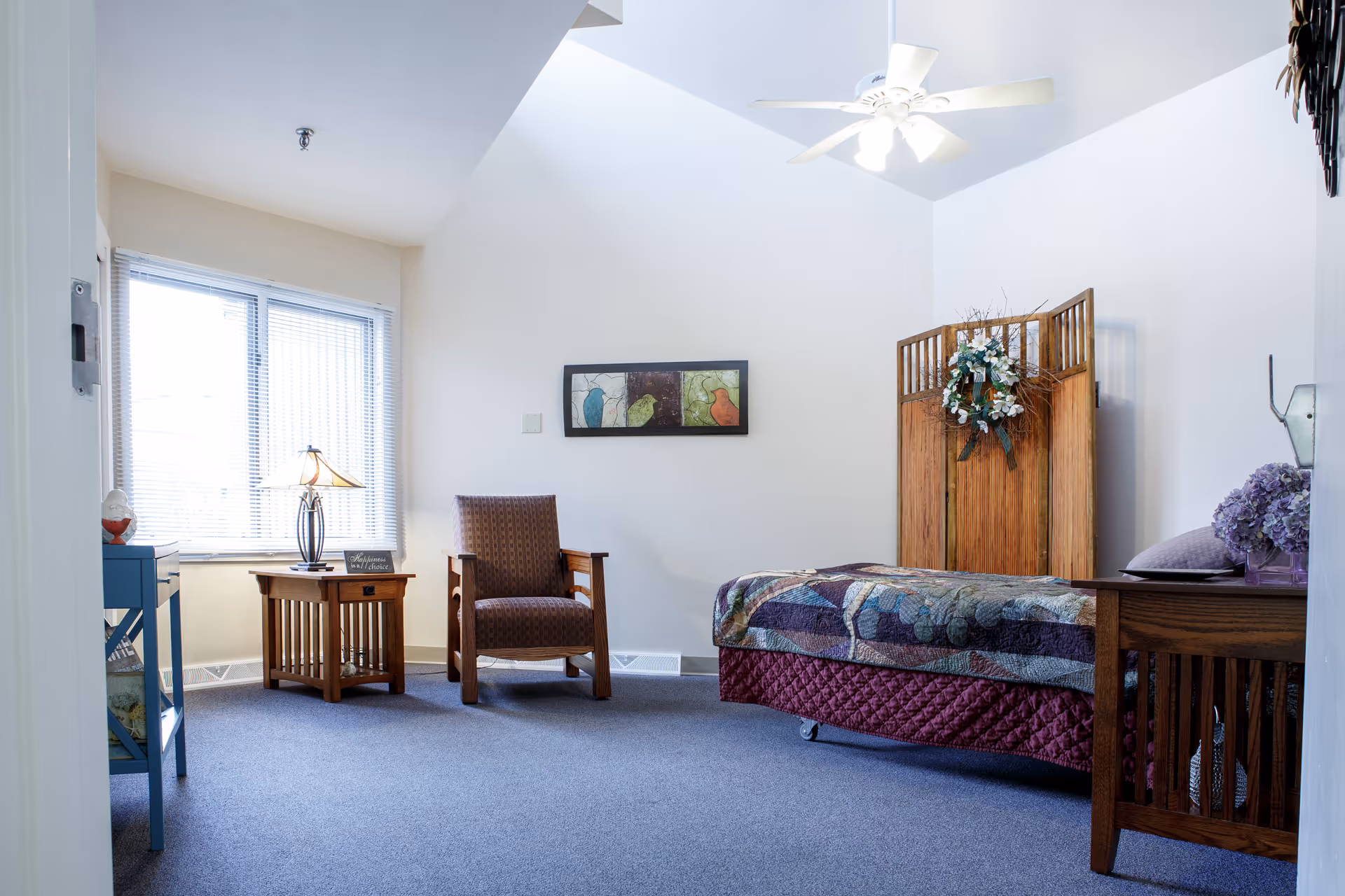 A bright bedroom with a single bed, wooden furniture, an armchair and a window with blinds.