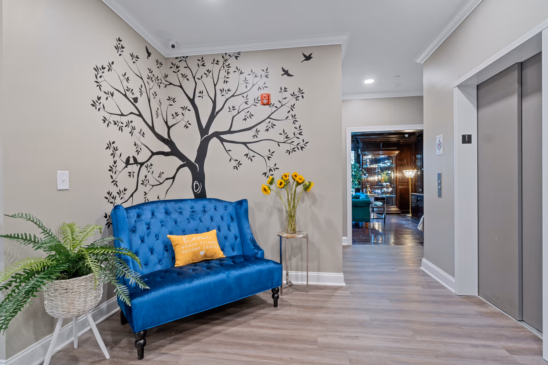 Lobby hallway with a blue tufted sofa beneath a tree wall decal, a potted plant and sunflowers, and elevator doors.
