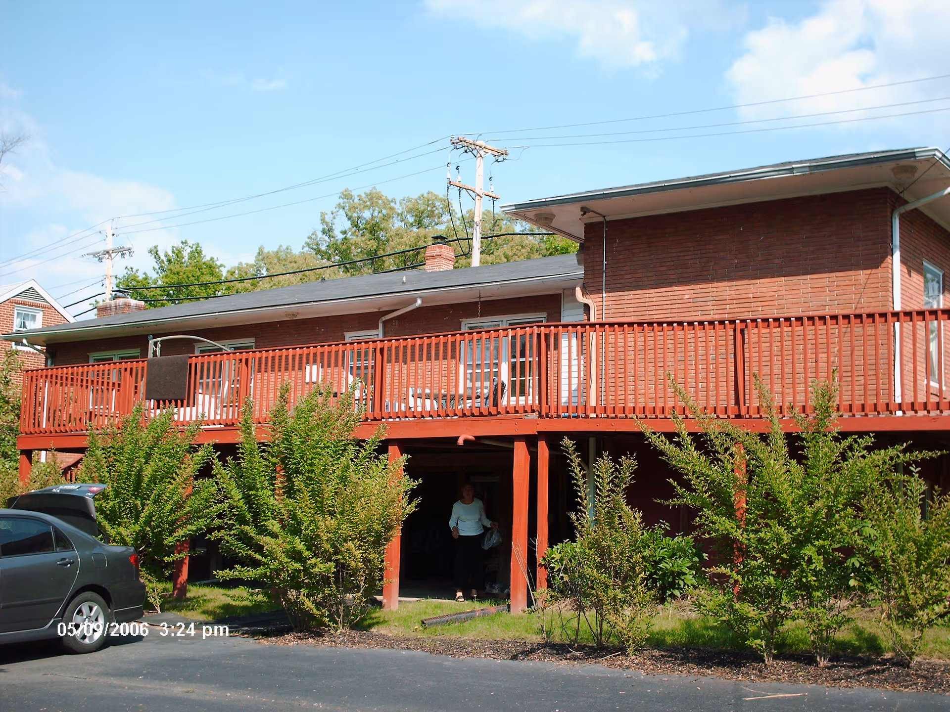 Two-story brick building with a long red front deck and surrounding shrubs; a car is parked nearby.