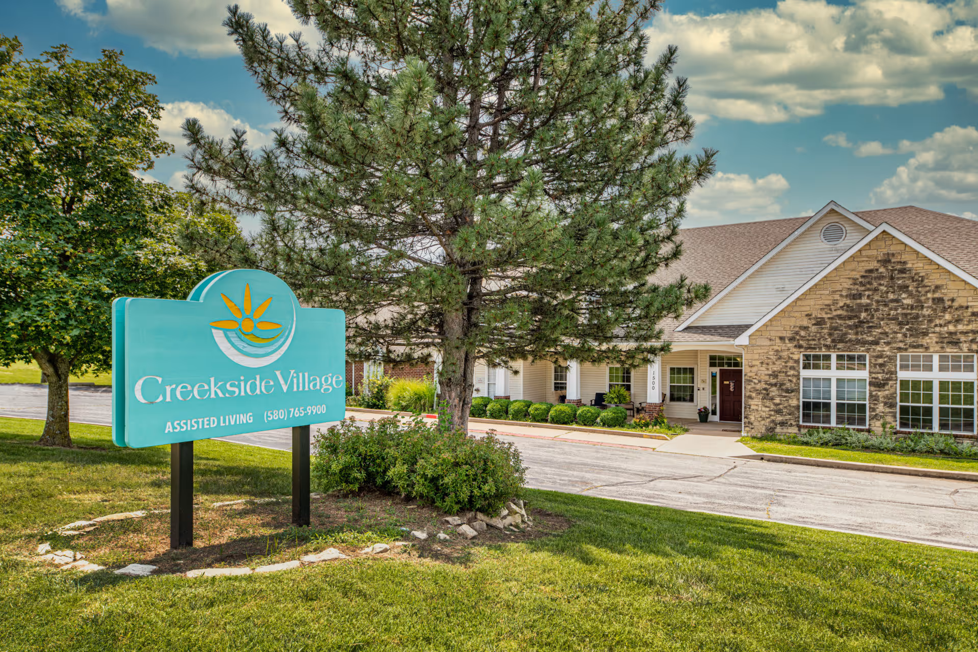 Exterior view of Creekside Village assisted living facility with a large green sign in the foreground displaying the facility name and phone number, surrounded by trees and grass, and a building with stone and siding in the background under a partly cloudy sky.