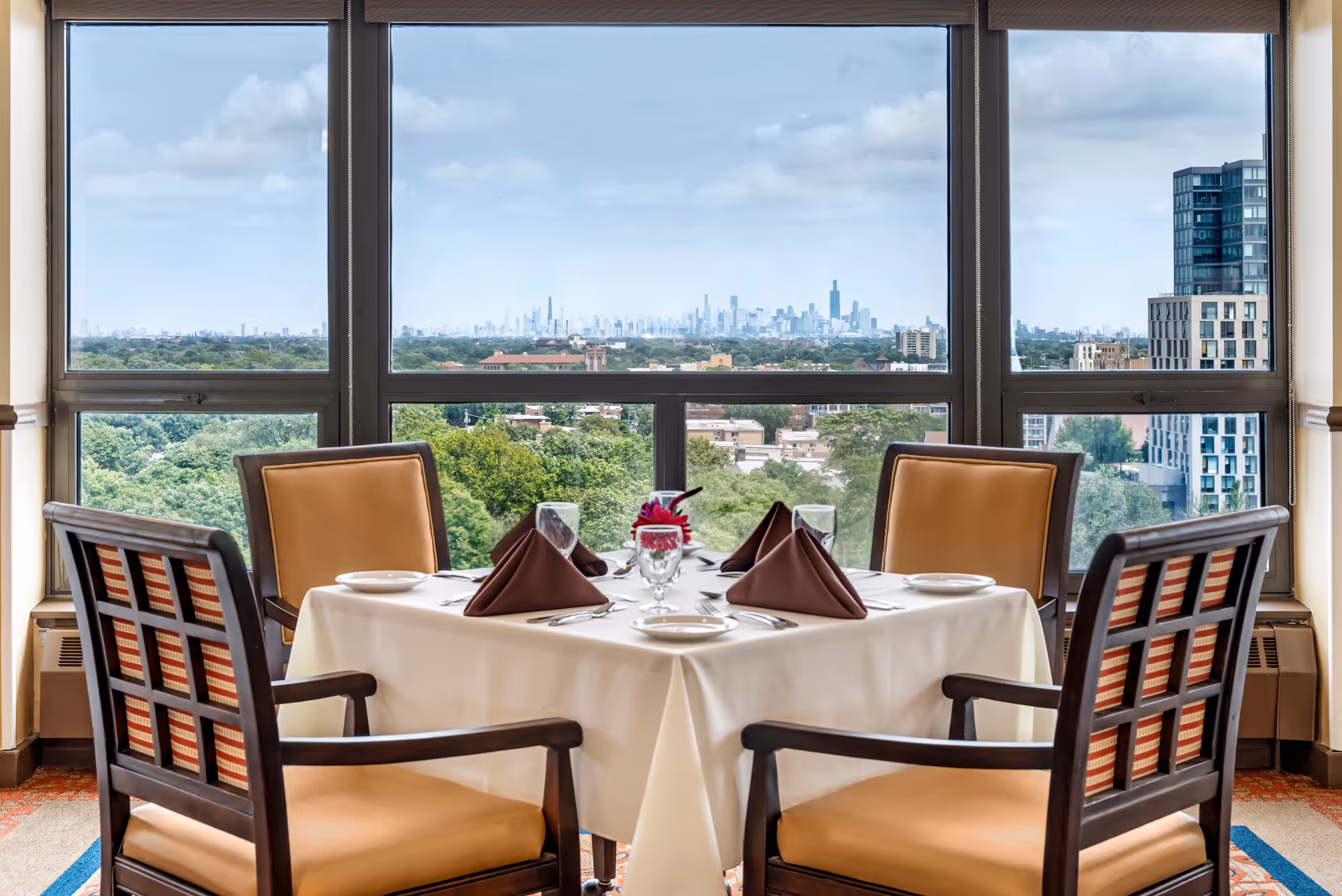 A neatly set dining table for four beside large windows with a view of trees and a distant city skyline.