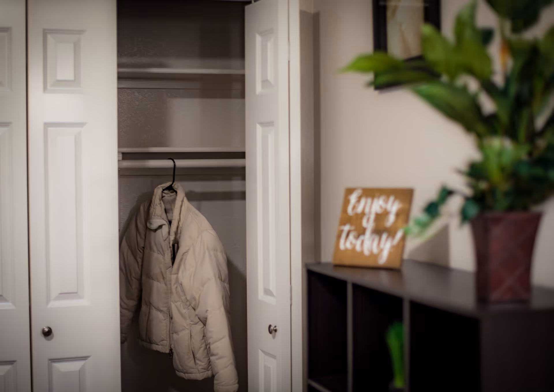 An open closet with white double doors showing a beige puffer jacket hanging inside. To the right, there is a dark wooden shelf with a small wooden sign that reads 'Enjoy today' and a potted plant with green leaves.