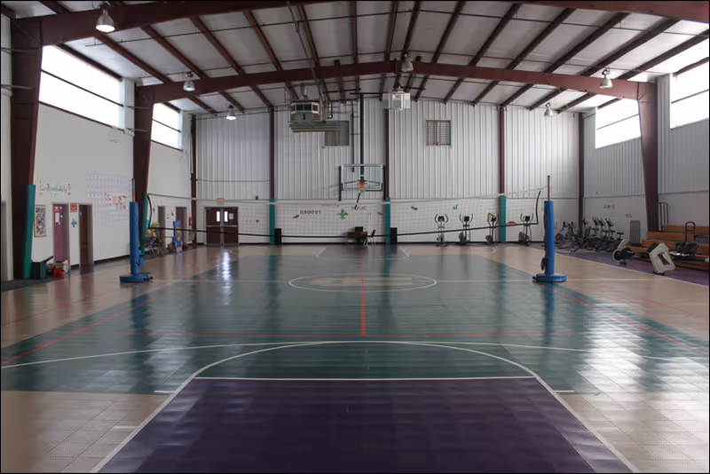 Indoor gymnasium with a basketball court and volleyball net set up. Exercise equipment is visible along the right wall, and the space has high ceilings with large windows letting in natural light.