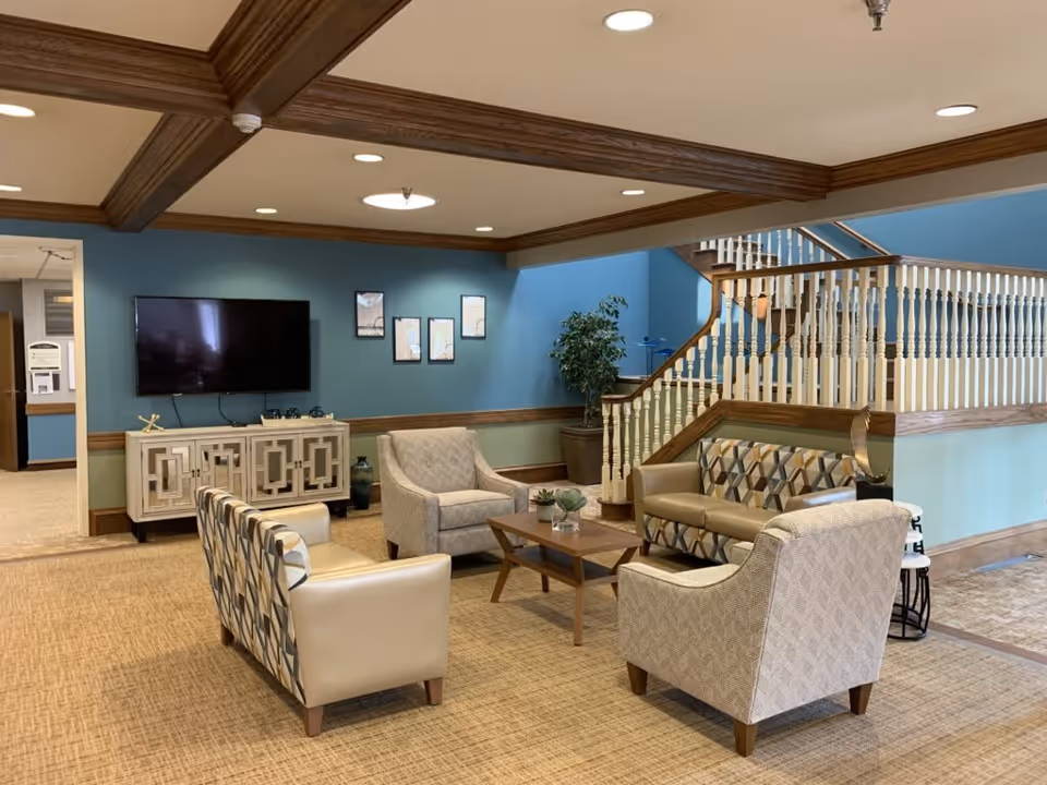 A cozy living room area in Westgate Assisted Living featuring a flat-screen TV mounted on a blue wall above a decorative cabinet. The seating arrangement includes two patterned armchairs, a beige armchair, and a beige loveseat with geometric patterns around a wooden coffee table. A staircase with white spindles and wooden handrails is visible in the background, along with a potted plant. The ceiling has wooden beams and recessed lighting.