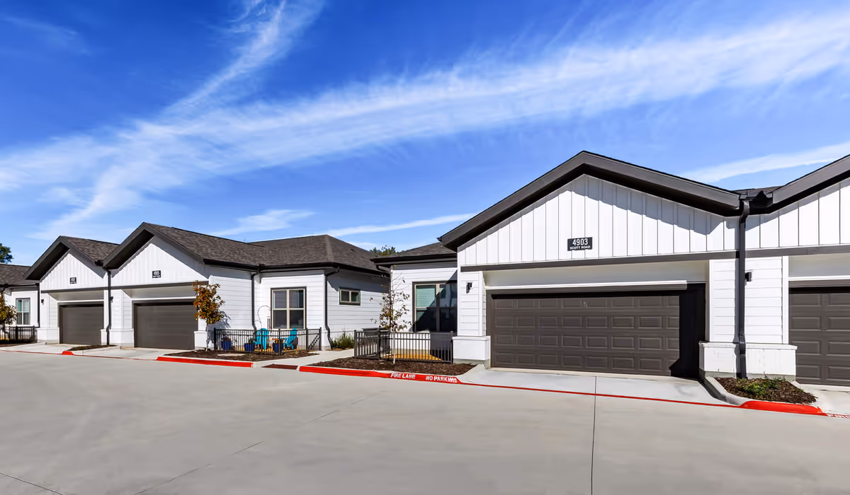 Exterior view of single-story residential units with white siding and dark brown garage doors under a blue sky with wispy clouds. Each unit has a small fenced patio area with outdoor chairs and young trees planted along the sidewalk.
