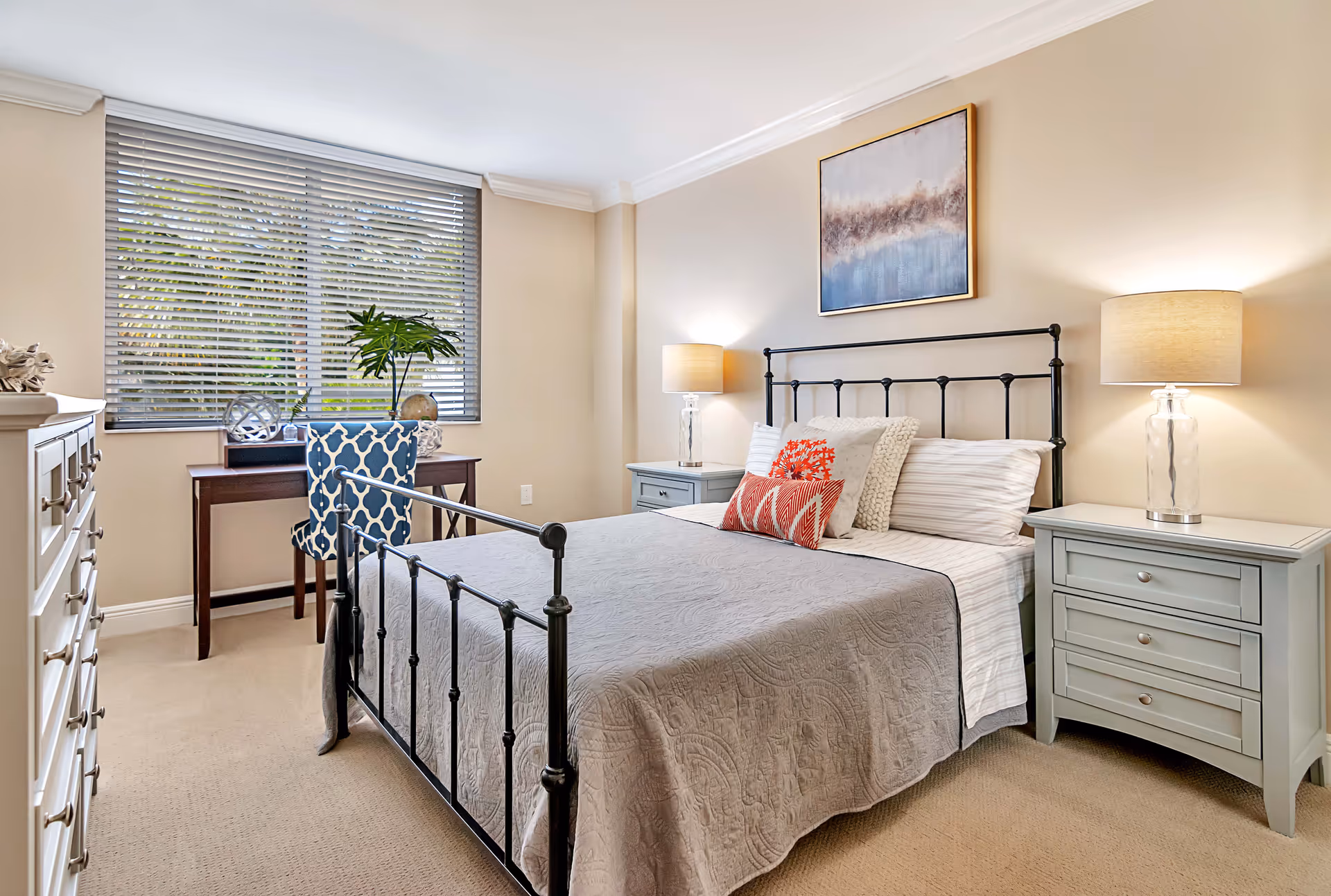 A neatly arranged bedroom with a black metal bed frame, gray quilt, and decorative pillows. Two matching light blue nightstands with lamps flank the bed. A wooden desk with a blue patterned chair is positioned in front of a window with blinds. A dresser is visible on the left side of the room, and a framed abstract painting hangs above the bed.