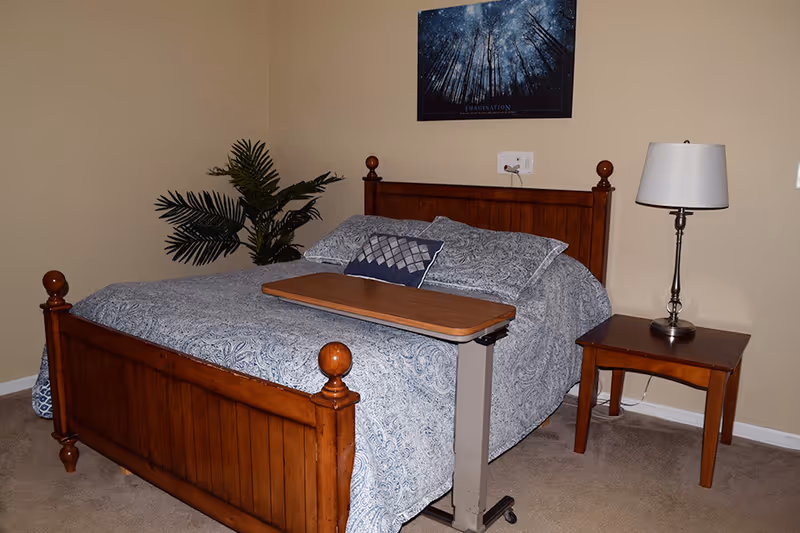 A bedroom with a wooden bed frame, blue patterned bedding, a small wooden side table with a lamp, a plant in the corner, and a framed picture of trees and stars on the wall.