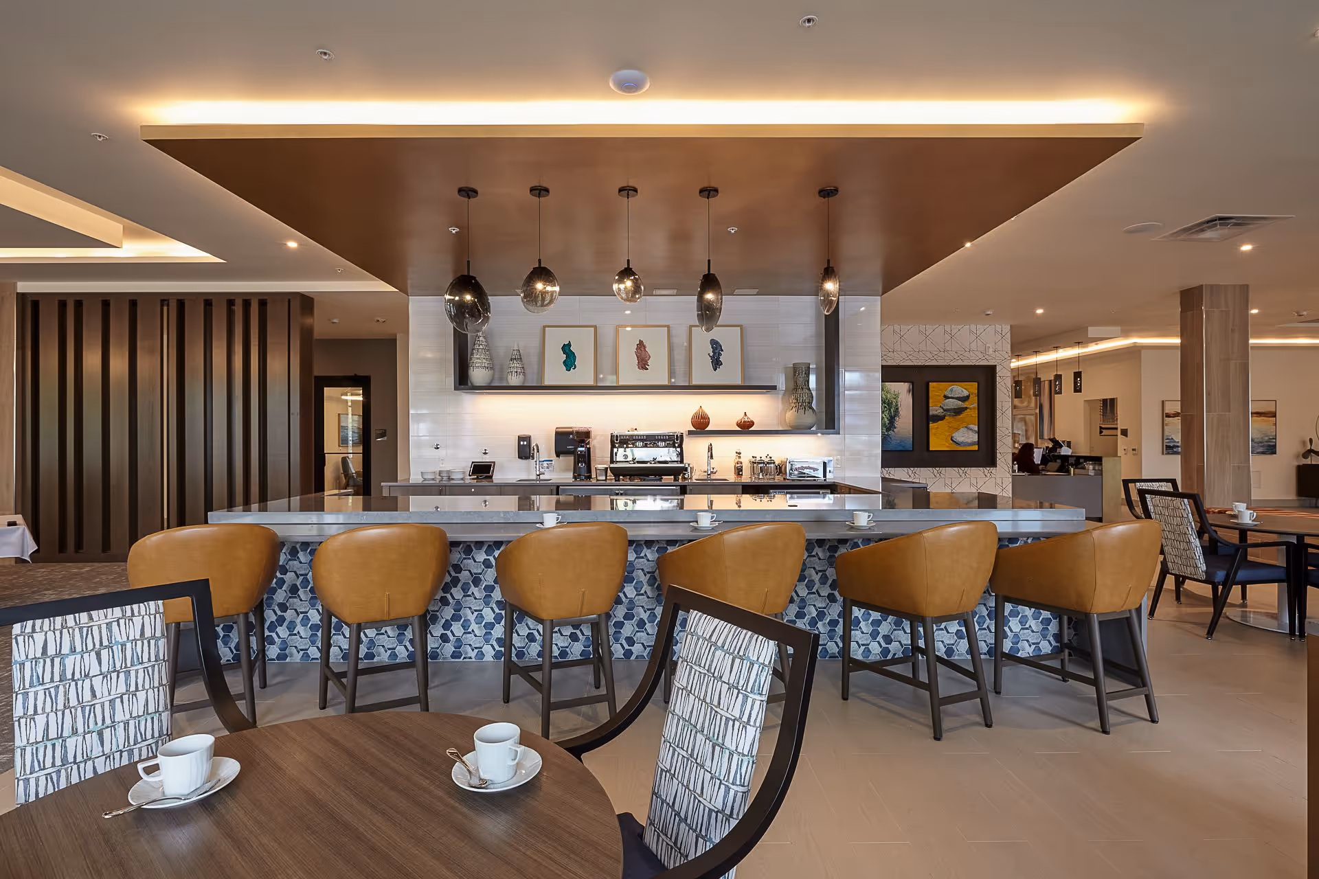 Modern senior living dining area featuring a central bar with tan leather stools, pendant lights, and nearby dining tables.