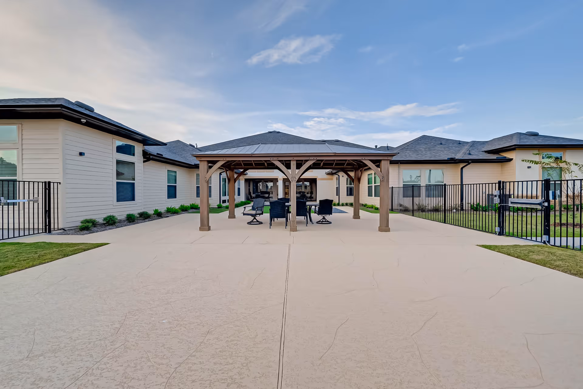 Outdoor courtyard with a central wooden pergola and seating between single-story assisted living buildings under a blue sky.