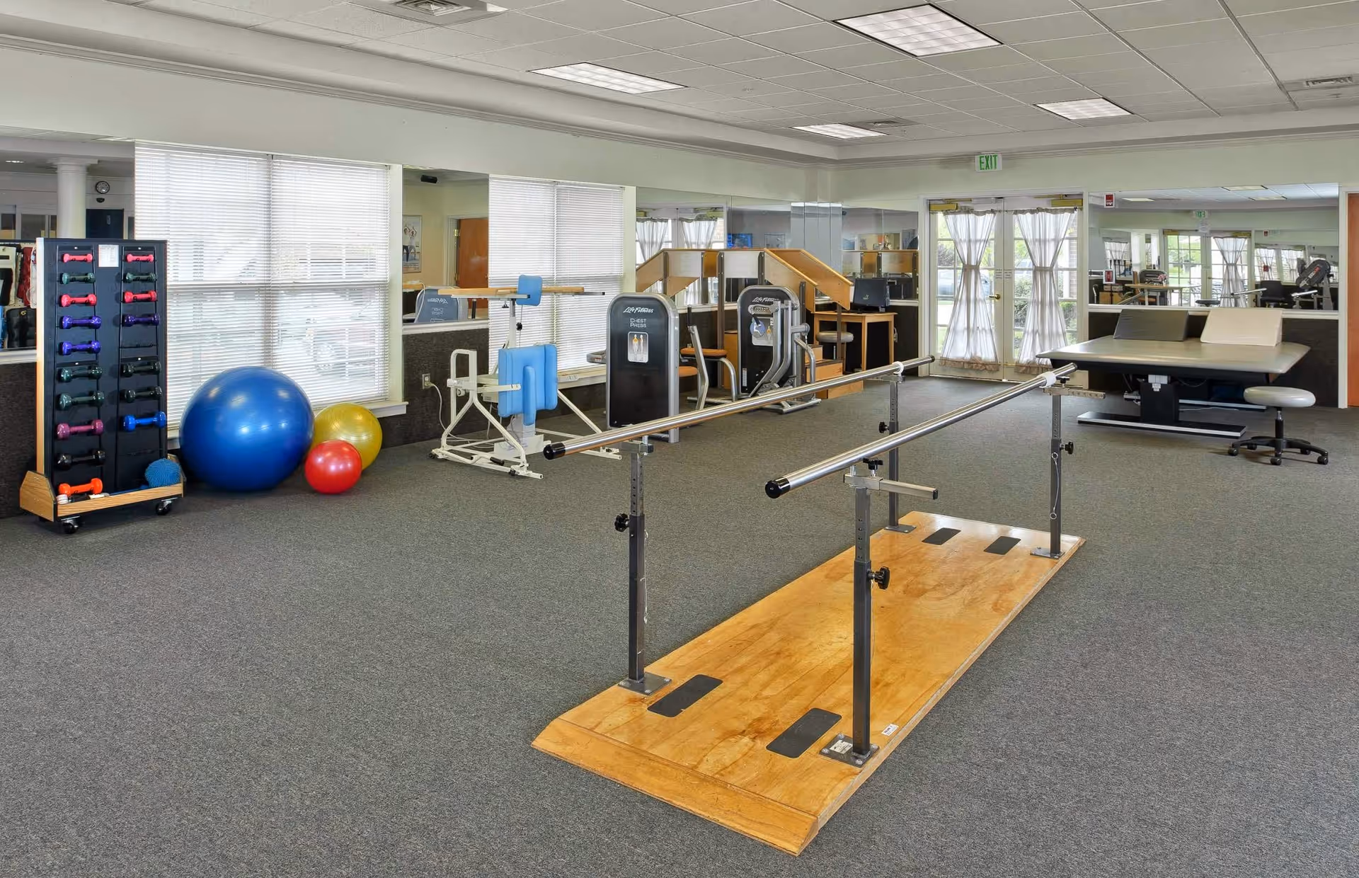 A spacious physical therapy or exercise room with parallel bars on a wooden platform in the center, various exercise equipment including a rack of colorful dumbbells, large exercise balls, and resistance machines. The room has large windows with blinds, carpeted floor, and a door with sheer curtains leading outside.