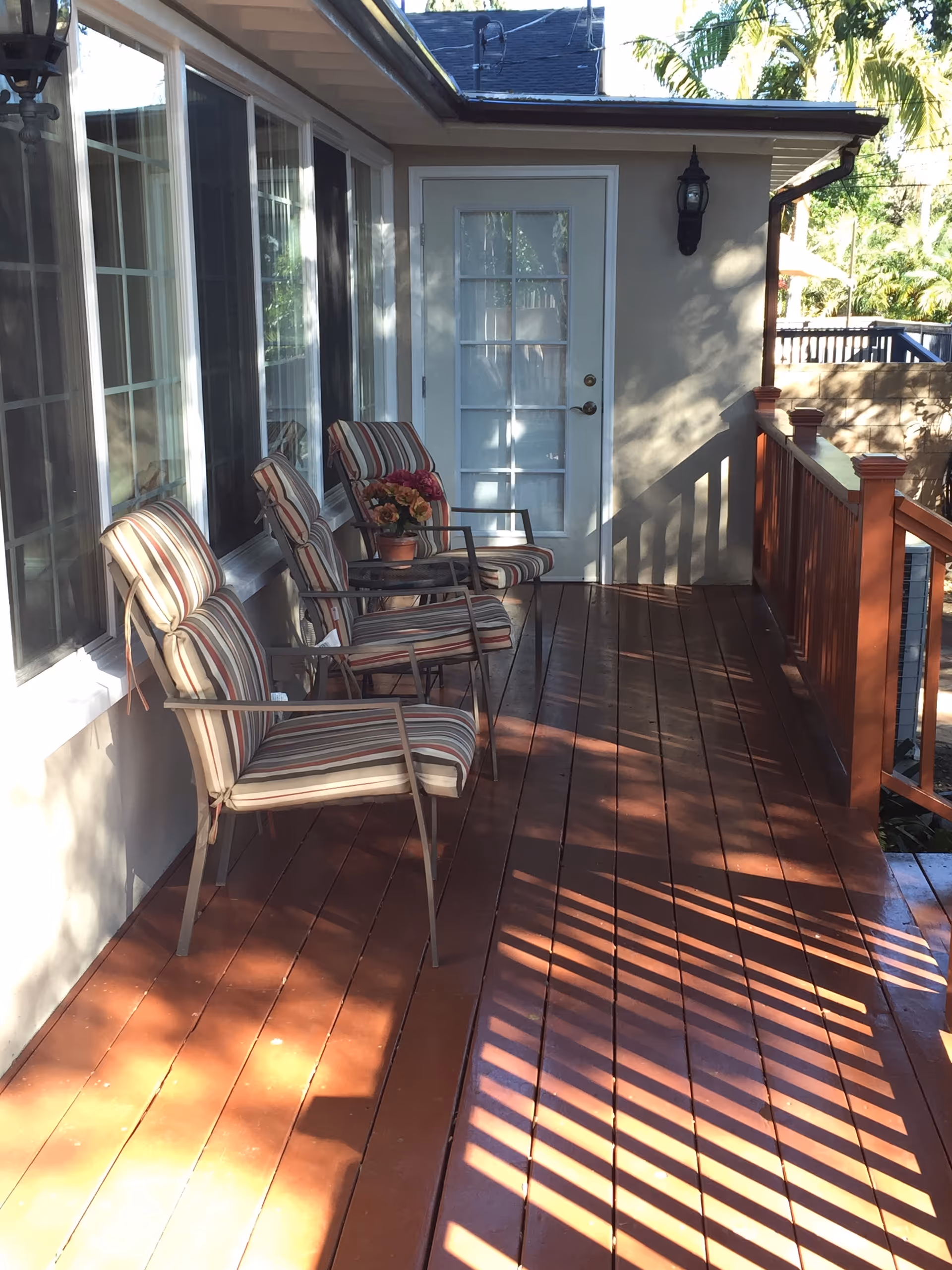 Sunlit wooden porch with striped cushioned chairs and a small table beside a glass-paneled door.