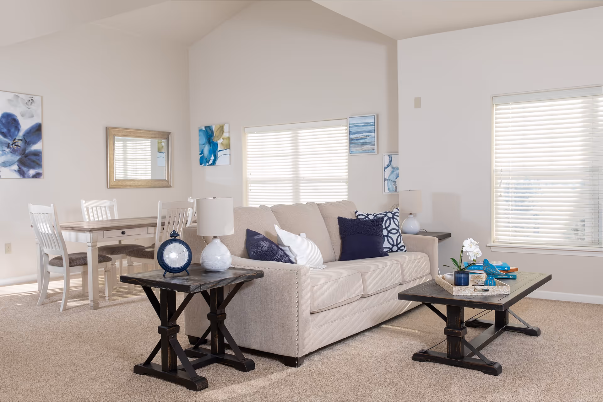 A bright living room with a beige sofa adorned with blue and white pillows, two dark wooden tables with decorative items including a lamp, clock, and a tray with books and a small plant. Behind the sofa is a dining area with a wooden table and four white chairs. The walls are decorated with blue-themed artwork and a mirror, and two windows with white blinds let in natural light.