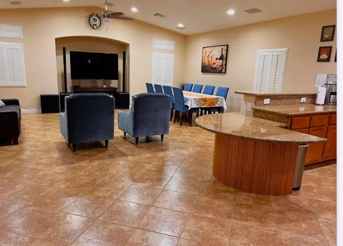A spacious living area with a tiled floor featuring two blue armchairs facing a large flat-screen TV mounted in a recessed wall niche. To the right, there is a dining table with blue chairs and a floral tablecloth. In the foreground, a kitchen island with a granite countertop and wooden cabinetry is visible. The walls are beige with white shutters on the windows and framed artwork hanging on the walls.