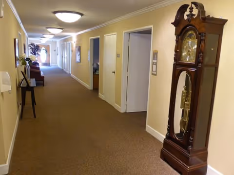 A long, carpeted hallway in a senior living facility with beige walls and ceiling lights. On the right side, there is a large wooden grandfather clock. Several doors line the hallway, and a small table with a flower arrangement is visible on the left side. At the far end, there is a window letting in natural light.