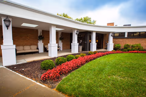 Curved covered entrance of a brick building with white columns, benches, glass doors, and a manicured lawn with red flower beds.