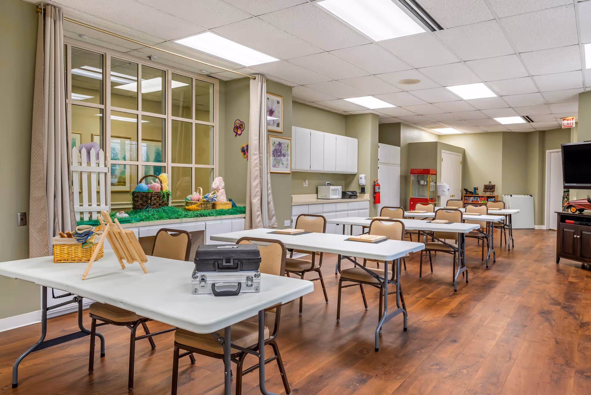 A well-lit activity room with several rectangular tables and chairs arranged in rows. The tables have some items on them including a small easel and cases. The room has light green walls, wood flooring, and a large window with a decorative display of baskets and Easter-themed decorations. There are white cabinets along one wall, a microwave, a refrigerator, and a popcorn machine in the background. A television is mounted on the right side of the room.
