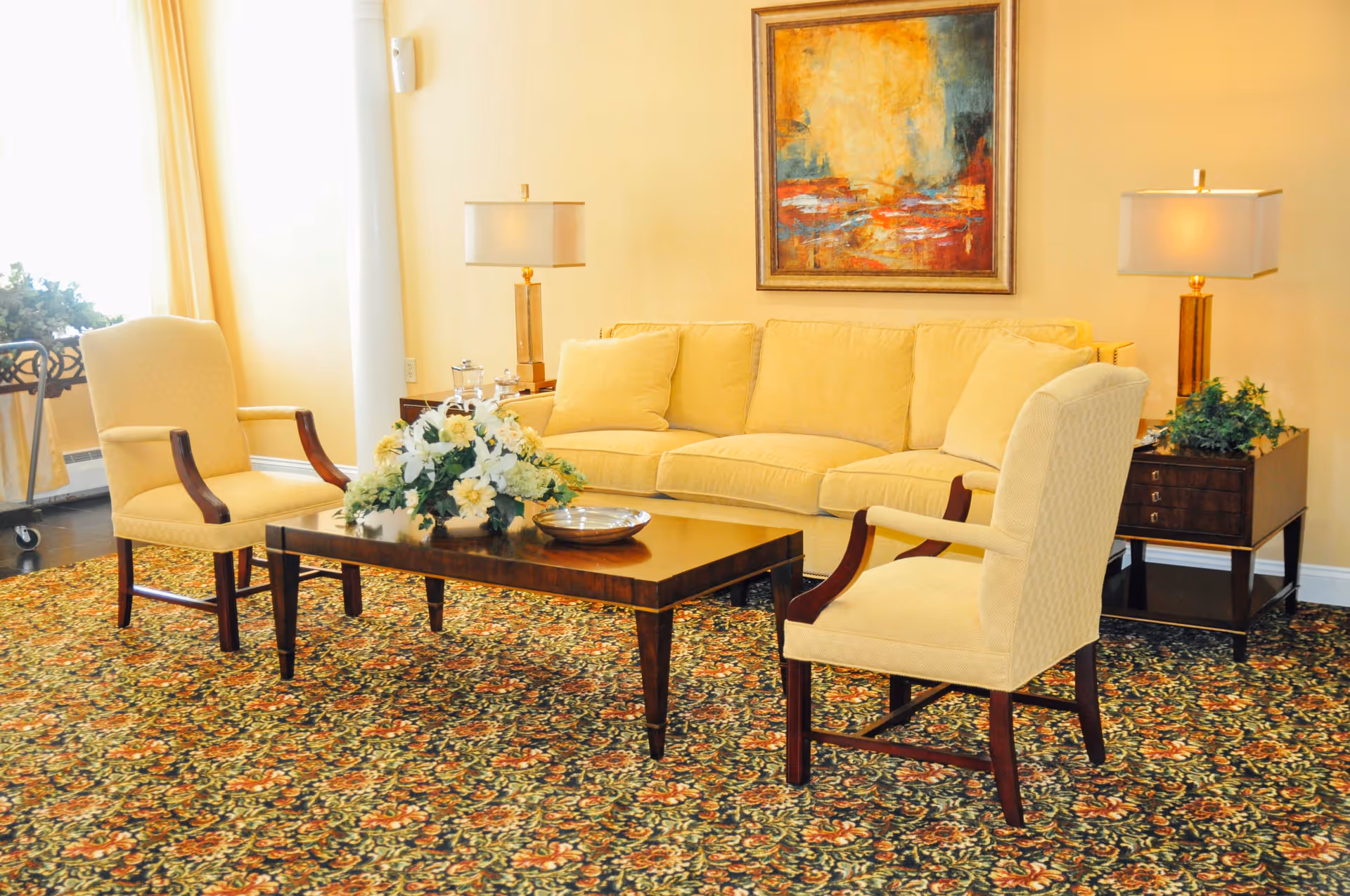 Cozy living room seating area with a beige sofa, two upholstered armchairs, a wooden coffee table topped with a floral arrangement, table lamps and framed artwork.