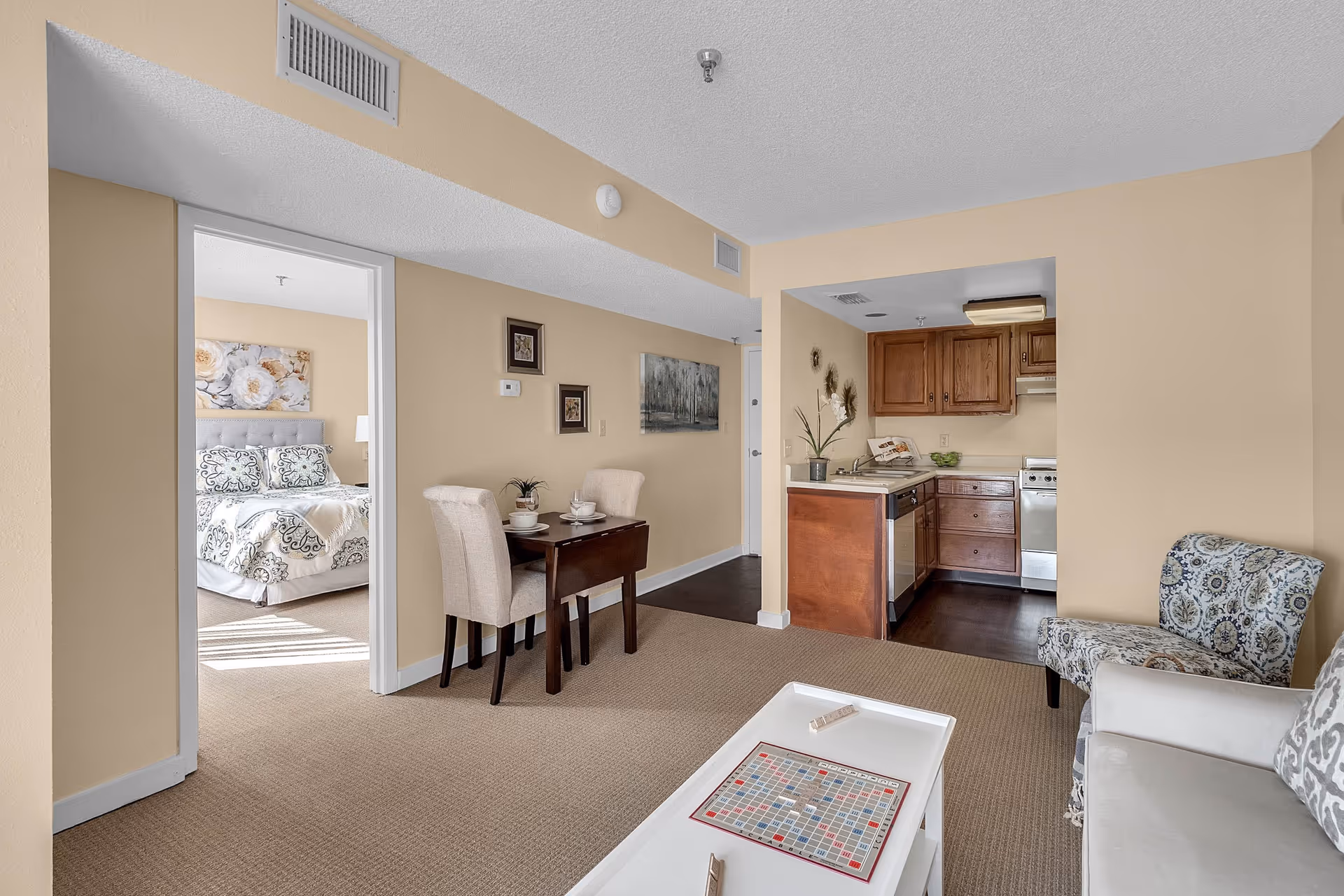 Interior view of a senior living facility apartment showing a small living area with a white coffee table featuring a Scrabble board game, a patterned armchair, and a white sofa. Adjacent to the living area is a compact kitchen with wooden cabinets and stainless steel appliances. A small dining table with two beige upholstered chairs is set against the wall. In the background, a bedroom with a bed adorned with floral bedding and a large flower artwork above the headboard is visible through an open doorway.