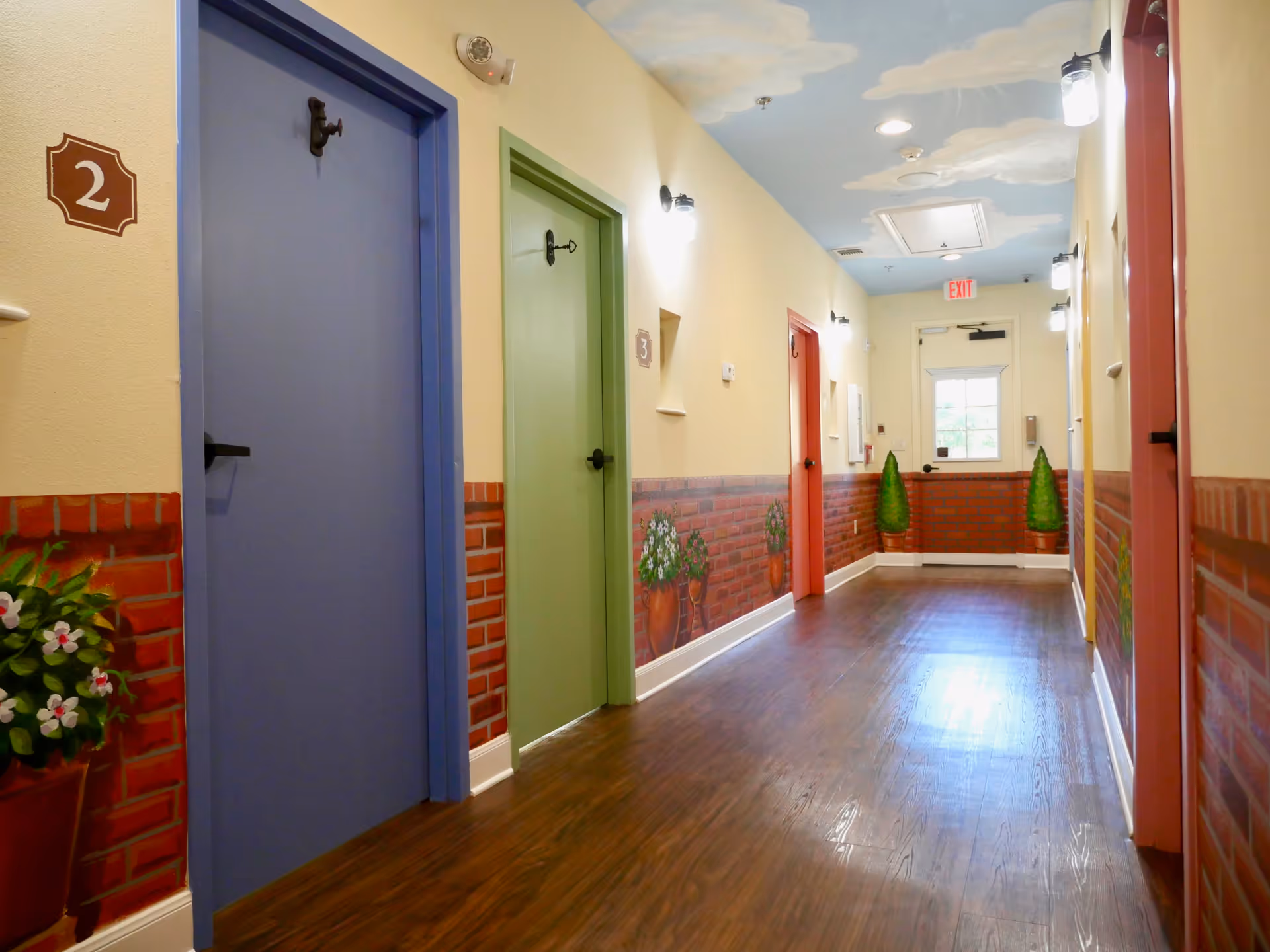 A brightly lit hallway in a senior living facility with colorful doors painted blue, green, and red, each numbered. The walls have a decorative brick pattern on the lower half with painted flower pots, and the ceiling is painted with clouds on a blue sky background. The floor is wooden, and there are wall-mounted lights along the corridor leading to an exit door with a window.