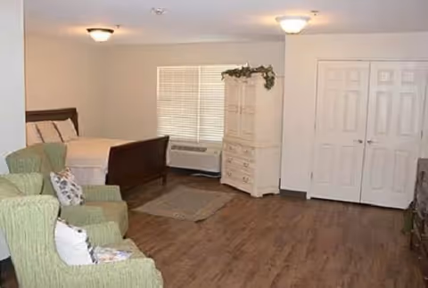 A senior living facility bedroom with a wooden bed, two green armchairs with pillows, a white dresser with decorative greenery on top, a window with blinds, a wall-mounted air conditioning unit, and a double-door closet. The room has wood flooring and neutral-colored walls.