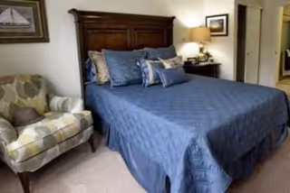 A bedroom with a large wooden headboard bed covered in a blue quilt and multiple pillows. To the left of the bed is a patterned armchair with a pillow. A nightstand with a lamp and framed pictures on the walls are visible in the background.