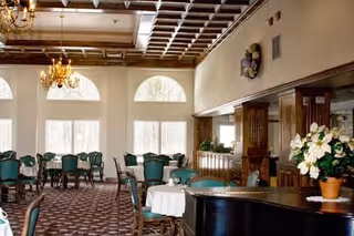 A spacious dining room with multiple tables covered with white tablecloths and surrounded by green upholstered chairs. The room features large arched windows letting in natural light, a patterned carpet, wooden paneling on the walls, and chandeliers hanging from the ceiling. There is a large floral arrangement on a dark wooden surface in the foreground.