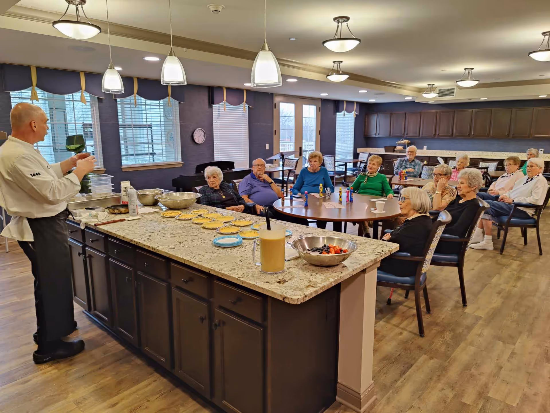 A chef is demonstrating or explaining something to a group of elderly people seated around a table and in chairs in a spacious room with a kitchen island in the foreground. The kitchen island has several plates with food items, a large bowl of fruit, and a drink. The room has large windows with blinds, pendant lights, and wooden flooring.