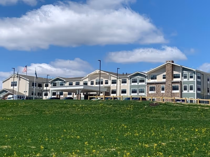 Front exterior of the Traditions of Columbus senior living building with a covered entrance, flagpoles, and a green lawn under a blue sky.