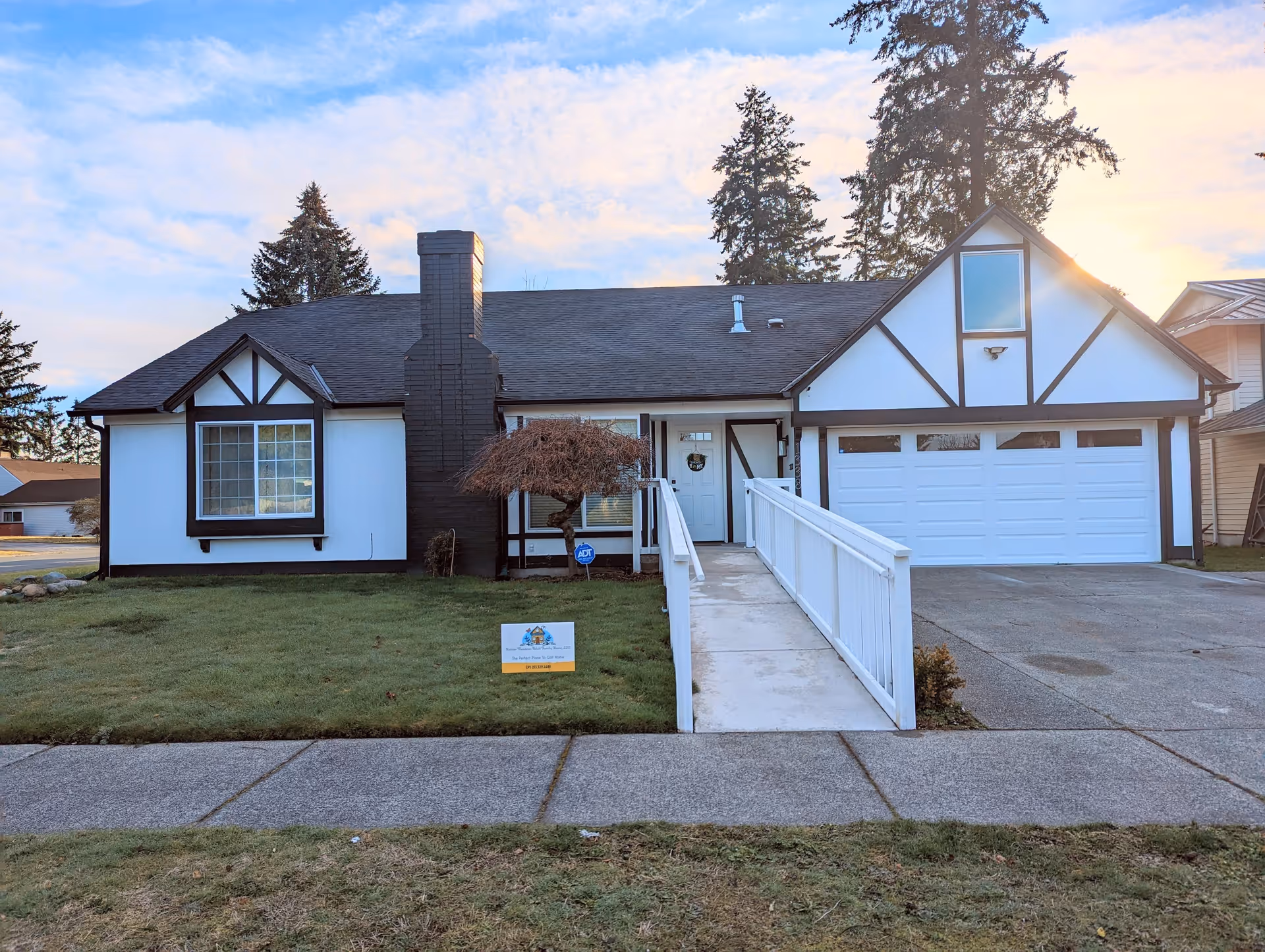Front exterior view of a single-story house with white walls and black trim, featuring a black chimney, a large window, a white front door with a wreath, a garage with a white door, and a concrete ramp leading to the entrance. The house is surrounded by a green lawn and tall trees in the background under a partly cloudy sky.