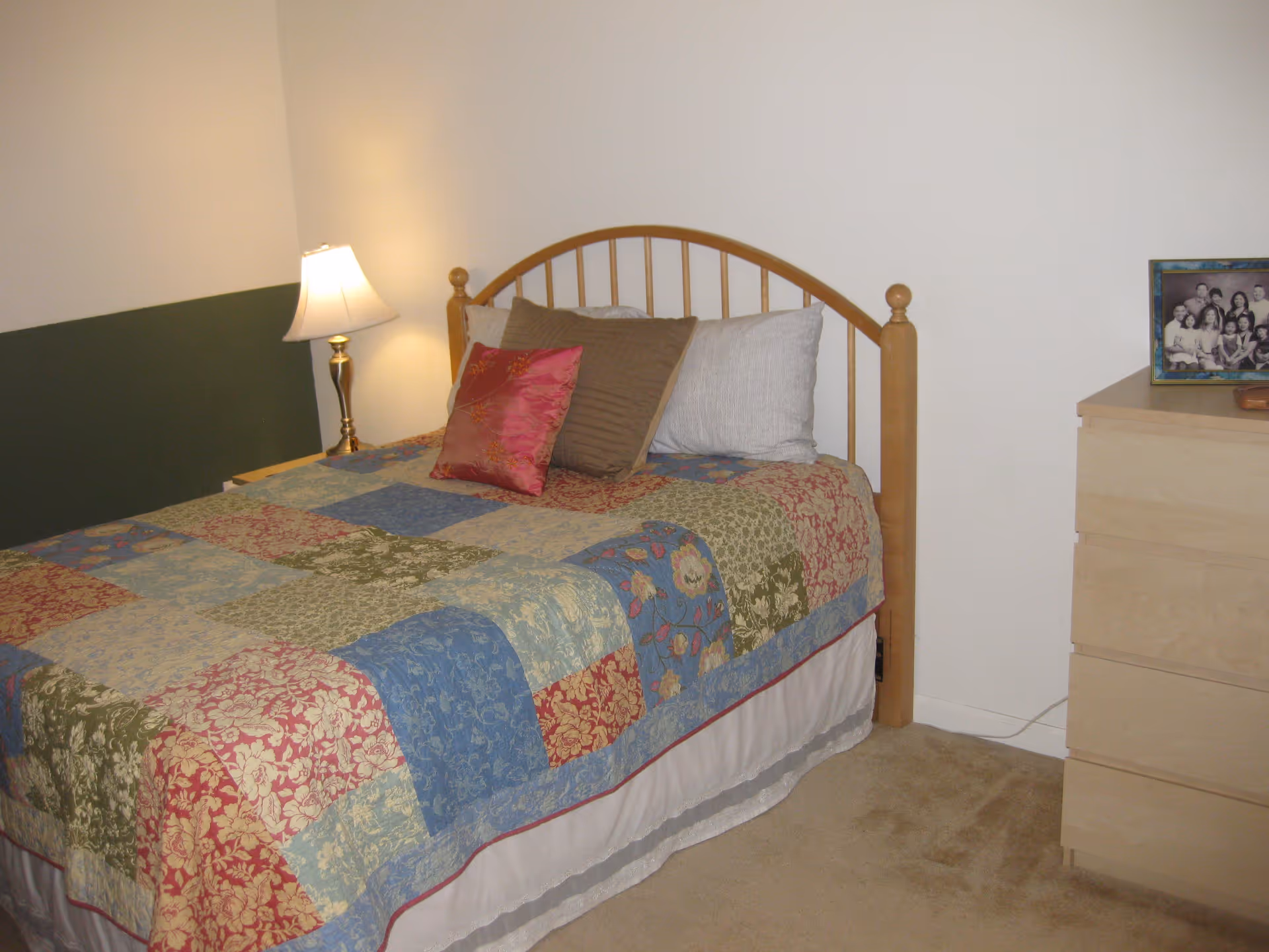 A bedroom with a wooden bed frame and a colorful patchwork quilt on the bed. There are three pillows on the bed, one white, one brown, and one red with a floral pattern. A bedside table with a lamp is next to the bed, and a light-colored wooden dresser with a framed family photo on top is visible on the right side. The walls are painted white with a dark green accent on one side.