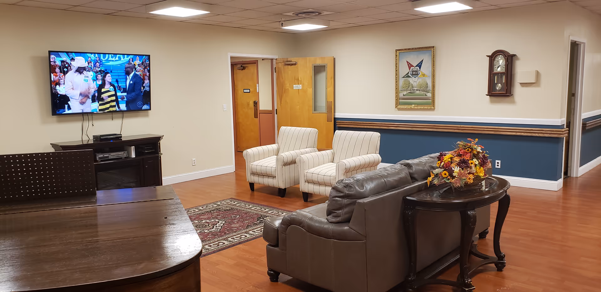 Community lounge area with a wall-mounted TV, couch and armchairs arranged on hardwood floor and a table with a floral arrangement.