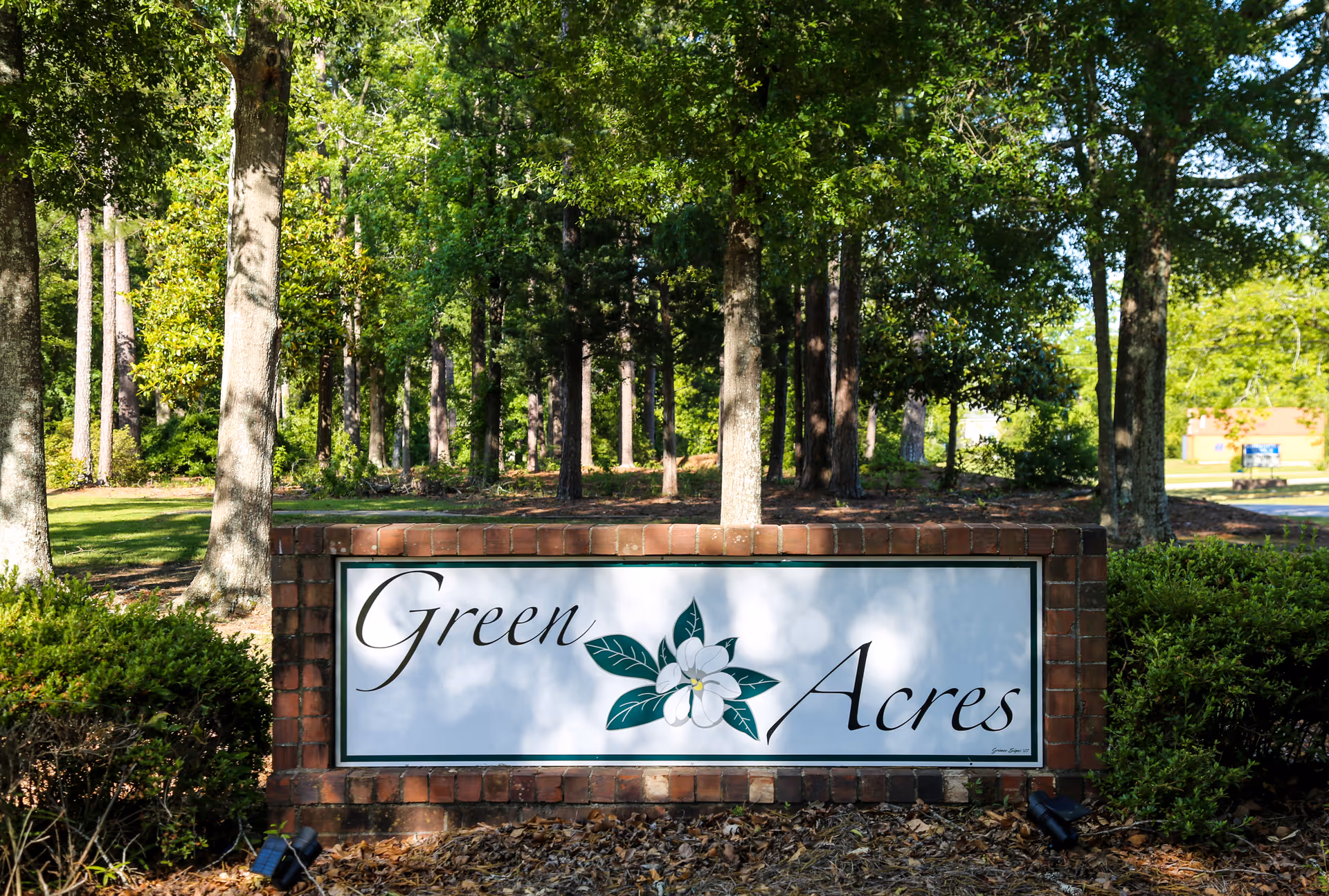 A brick sign with a white panel displaying the text 'Green Acres' with a flower graphic in the center, surrounded by green trees and bushes in an outdoor setting.