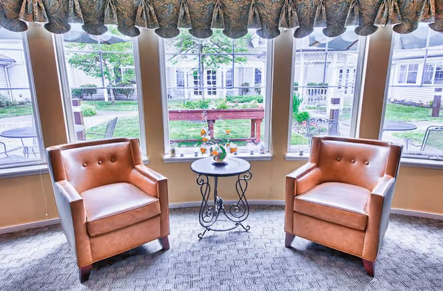 Two brown leather armchairs positioned on either side of a small round metal table with a flower vase, set in front of large windows with a view of a green outdoor courtyard with patio furniture and buildings.