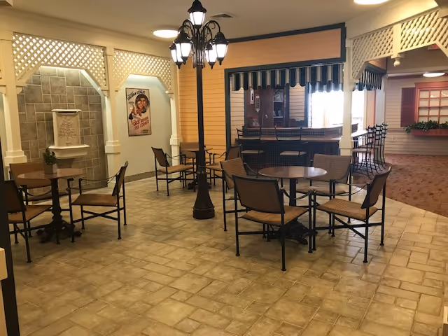 Indoor seating area with several round tables and chairs arranged on a tiled floor. A decorative streetlamp-style light fixture stands in the middle. The walls have a lattice design and a small water fountain is mounted on one wall. There is a counter with bar stools under a striped awning, and a vintage-style poster is visible on the wall.