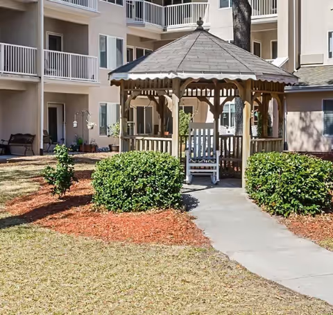 Wooden gazebo with a white rocking chair in a landscaped courtyard in front of a multi-story residential building.