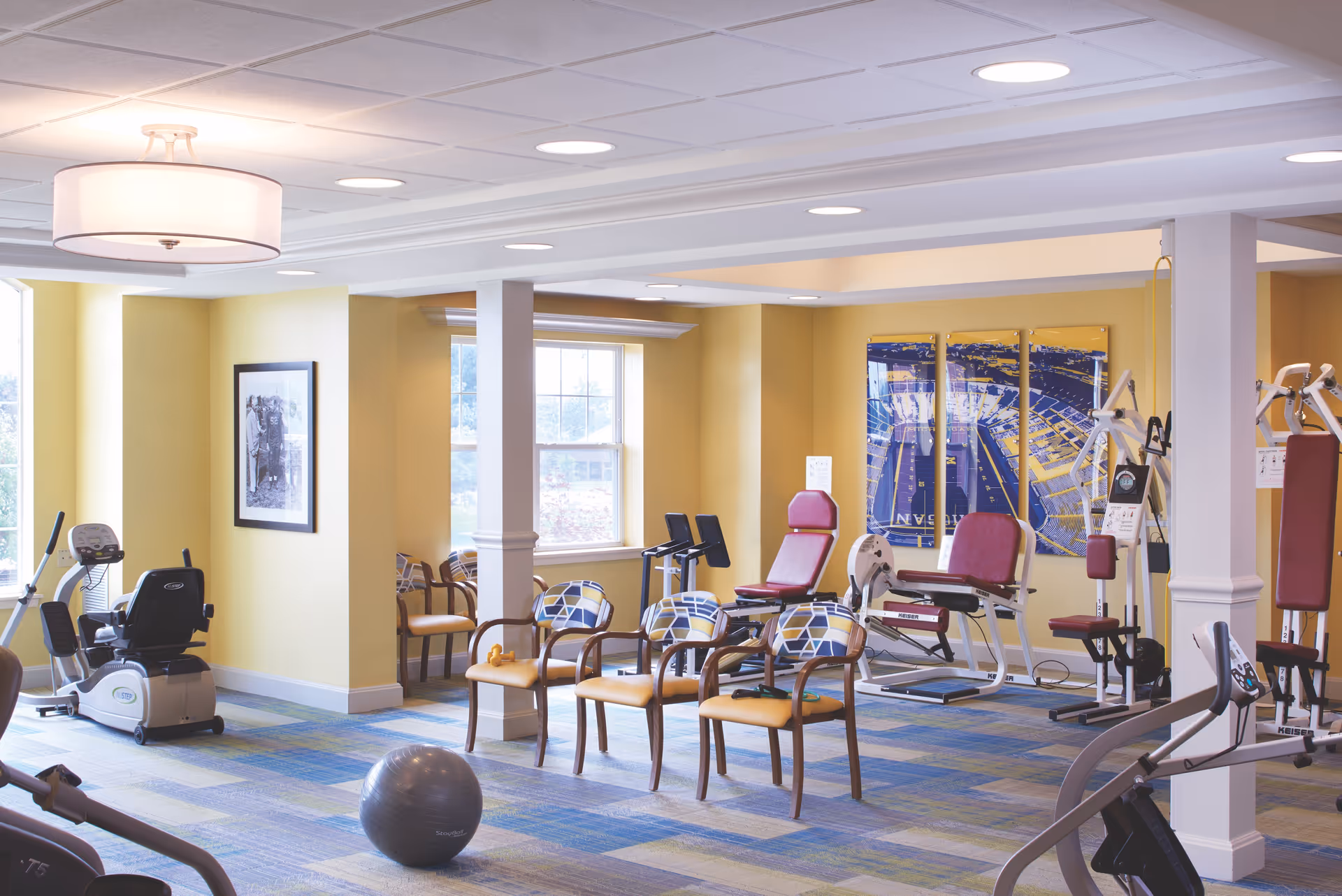 A bright exercise room in a senior living facility with yellow walls and large windows. The room contains various exercise equipment including stationary bikes, weight machines, and a stability ball. There are several chairs with patterned cushions arranged in the center of the room. The floor is carpeted with a blue and green pattern, and there are framed pictures and artwork on the walls.