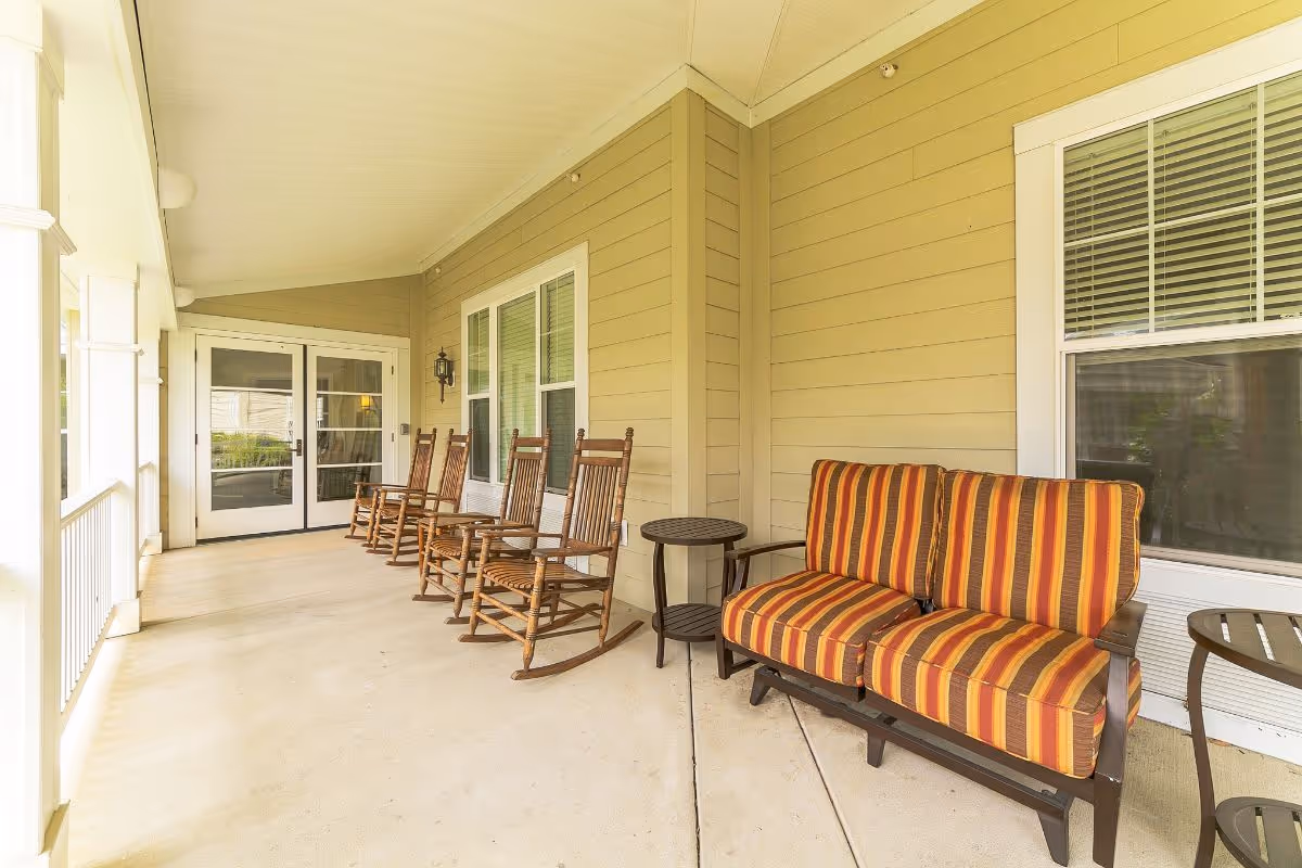 Covered outdoor porch area with four wooden rocking chairs lined up against a beige wall, two small round tables, and a cushioned bench with striped orange, yellow, and brown upholstery. White framed windows and double glass doors are visible in the background.