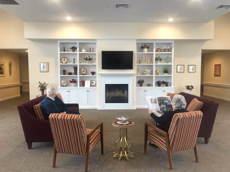 A cozy common area in a senior living facility with two elderly people seated on striped armchairs facing each other. Behind them are two purple sofas, a fireplace with a mounted TV above it, and built-in shelves decorated with various vases, plants, and decorative items. The room is softly lit with recessed ceiling lights.