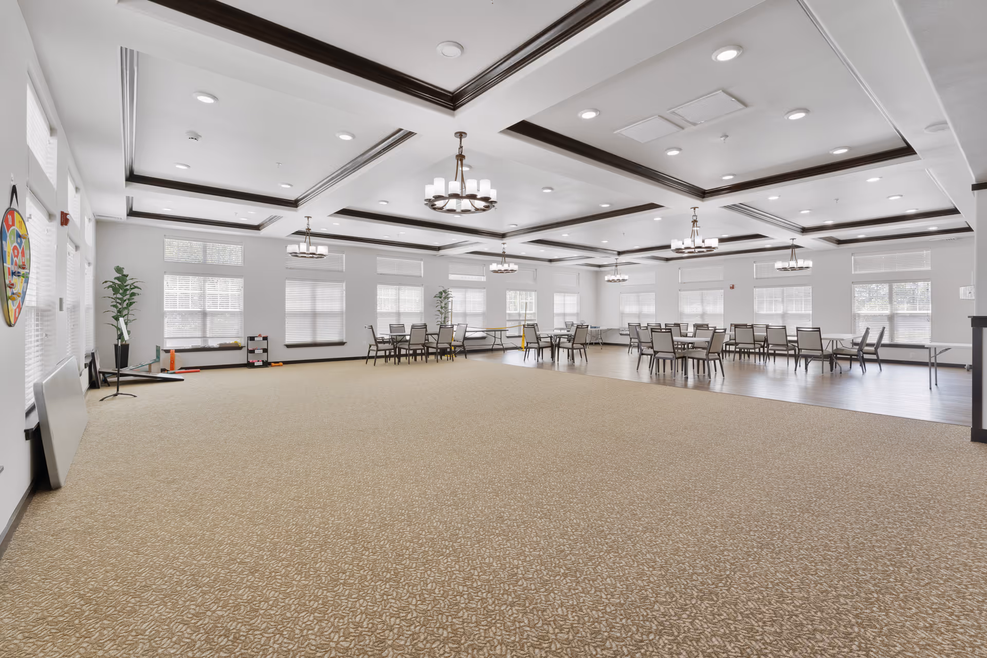 A spacious, well-lit common area with beige patterned carpet and wood flooring. Several tables and chairs are arranged near the windows, and there are large windows with blinds letting in natural light. The ceiling features recessed lighting and decorative dark wood beams with hanging chandeliers. A dartboard and some game equipment are visible on the left side of the room.