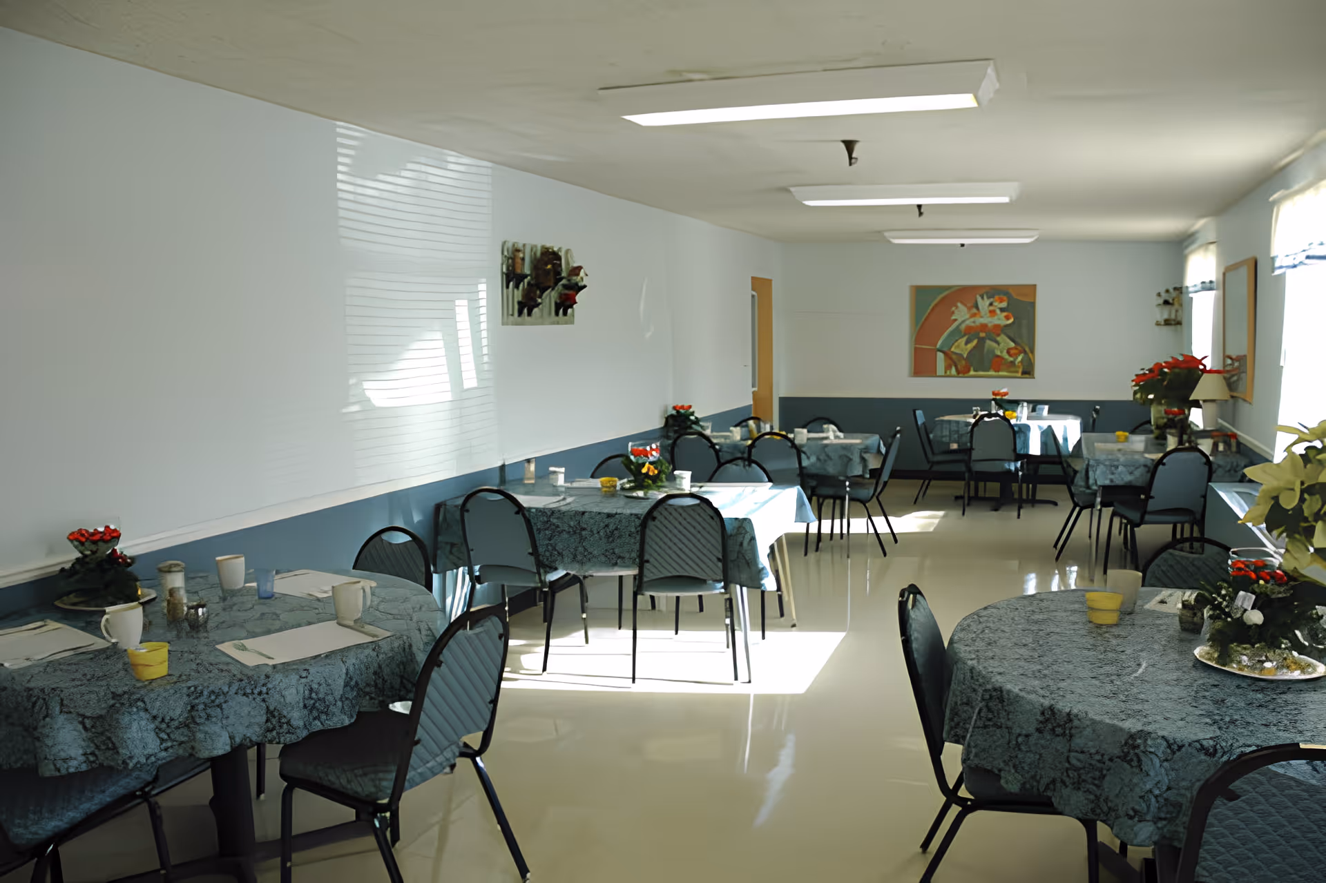 A dining room with several round tables covered with blue patterned tablecloths. Each table has chairs around it and is set with cups, napkins, and small flower arrangements. The room has light blue walls, a shiny floor, fluorescent ceiling lights, and windows on the right side letting in natural light. There is artwork on the far wall and additional plants and decorations on the windowsills.