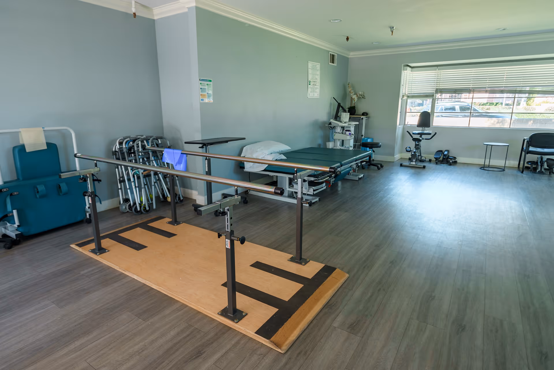 A bright rehabilitation room with parallel walking bars, therapy beds, walkers, and exercise equipment near a large window.