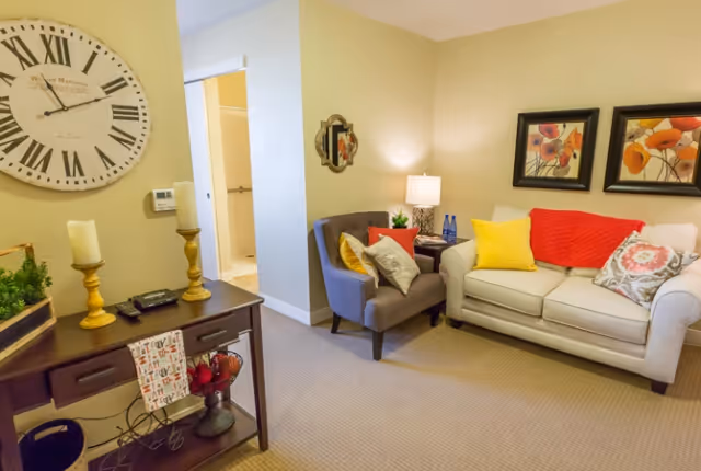 Well-lit living room with a sofa, armchair, console table, large wall clock, and framed floral artwork.