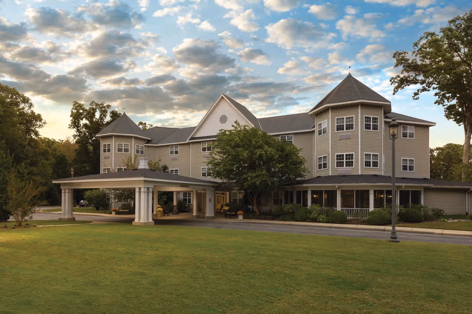 Exterior view of Mennowood Retirement Community building with a covered entrance, surrounded by green lawn and trees under a partly cloudy sky.