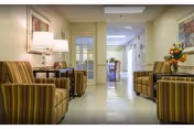 A hallway in a senior living facility with striped armchairs arranged along the walls, a side table with a lamp, framed artwork on the walls, and a flower arrangement on a table. The hallway leads to a brighter area with more seating visible in the distance.