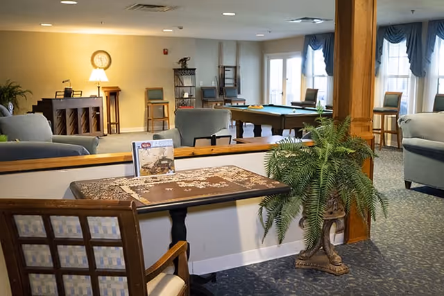 A spacious senior living common area with comfortable seating, a table with a partially completed puzzle, a large potted plant, a piano, a clock on the wall, and a pool table near windows with blue curtains.