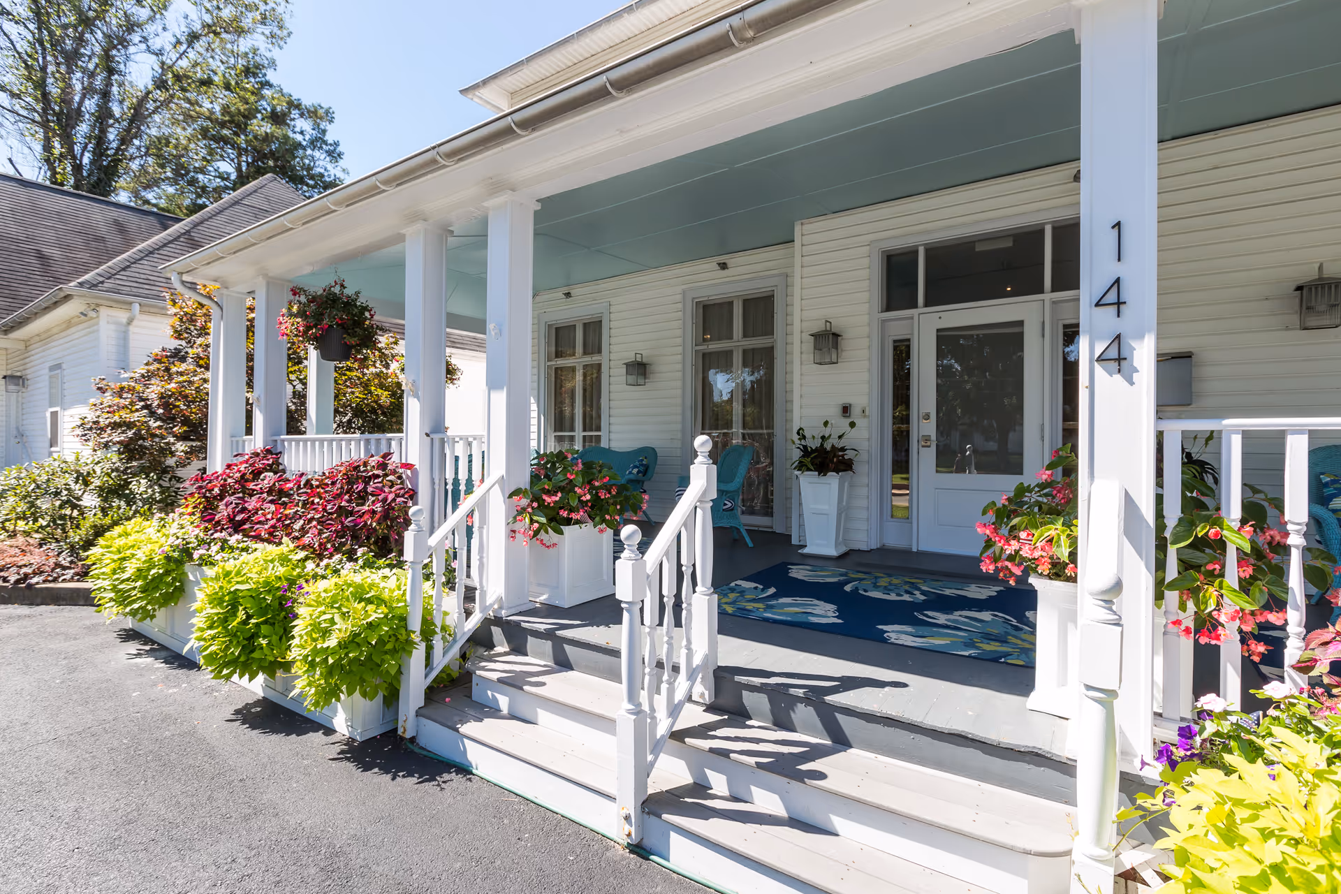 Front porch of a white building with the number 144 on a column, decorated with potted plants and flowers, two blue chairs, and a blue floral rug on the porch floor.