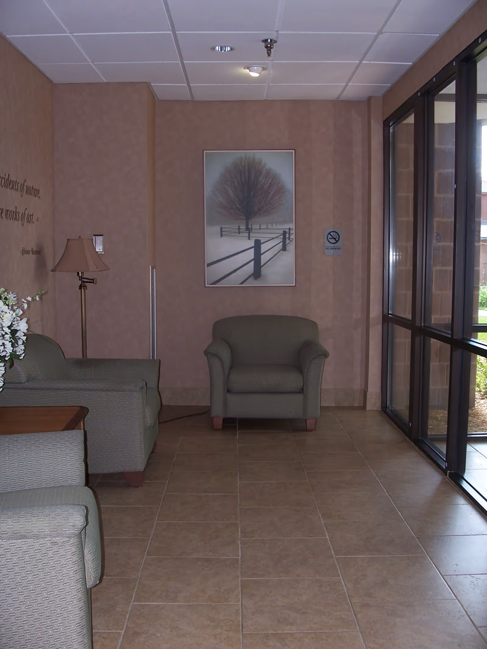 A small seating area in a facility with three green upholstered armchairs, a floor lamp, a wooden side table with a flower arrangement, and a large window wall letting in natural light. A framed picture of a tree and fence in a snowy landscape hangs on the wall, along with a no smoking sign.
