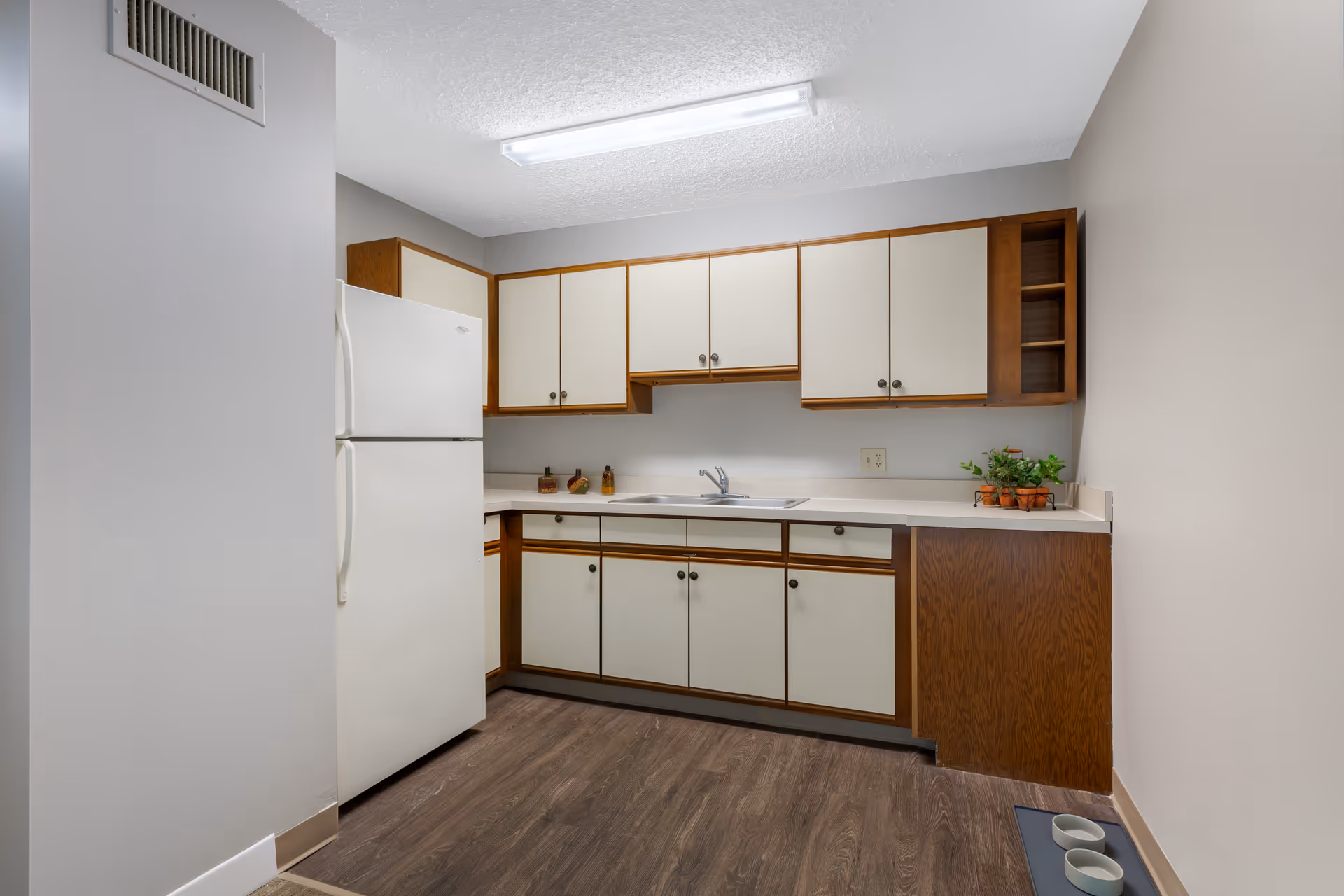 A small kitchen with wood and cream-colored cabinets, a white refrigerator, a double sink, and a countertop with small decorative items and potted plants. The floor is wood-patterned, and there are two empty pet bowls on a mat in the corner.
