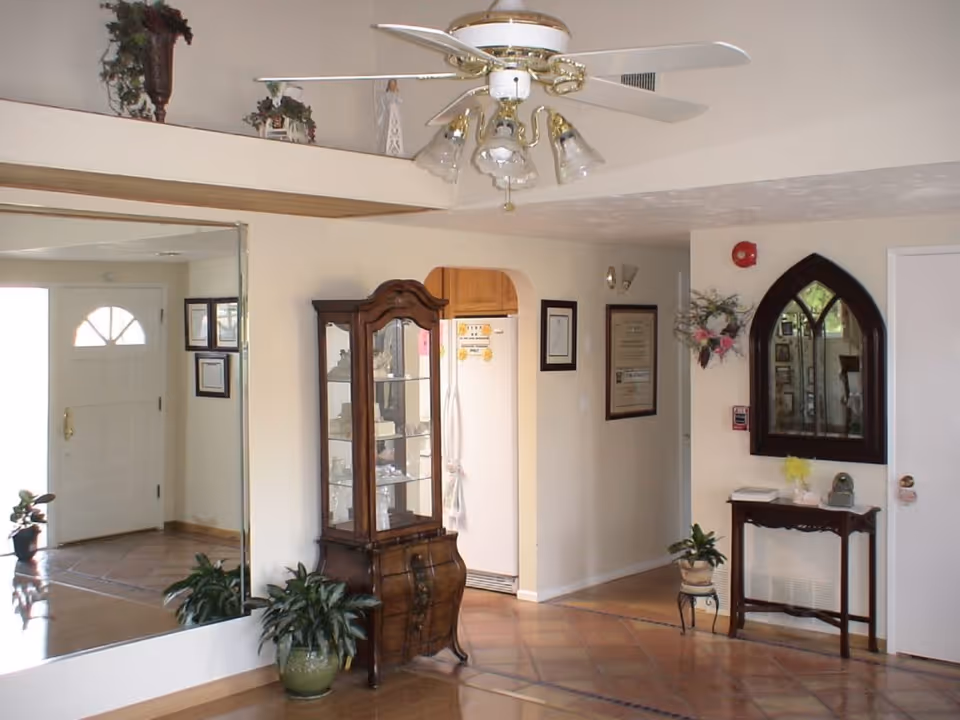 Interior view of a living area with a white ceiling fan with lights, a wooden display cabinet with glass doors, a large wall mirror reflecting the entrance door, framed certificates on the wall, a small wooden table with a decorative mirror above it, and several potted plants on the floor.
