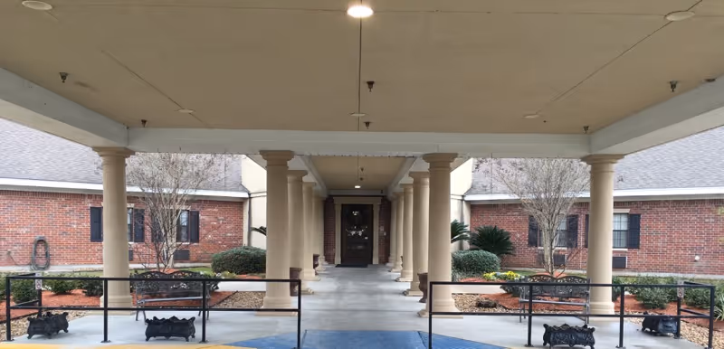 Covered entrance portico with columns leading to the main double doors of a brick senior living facility, flanked by landscaped beds and benches.